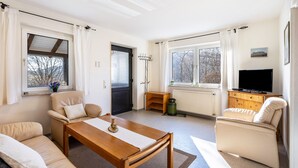 Ein Wohnzimmer mit Sofa, Sessel, Holztisch, Fernseher und Fenstern.