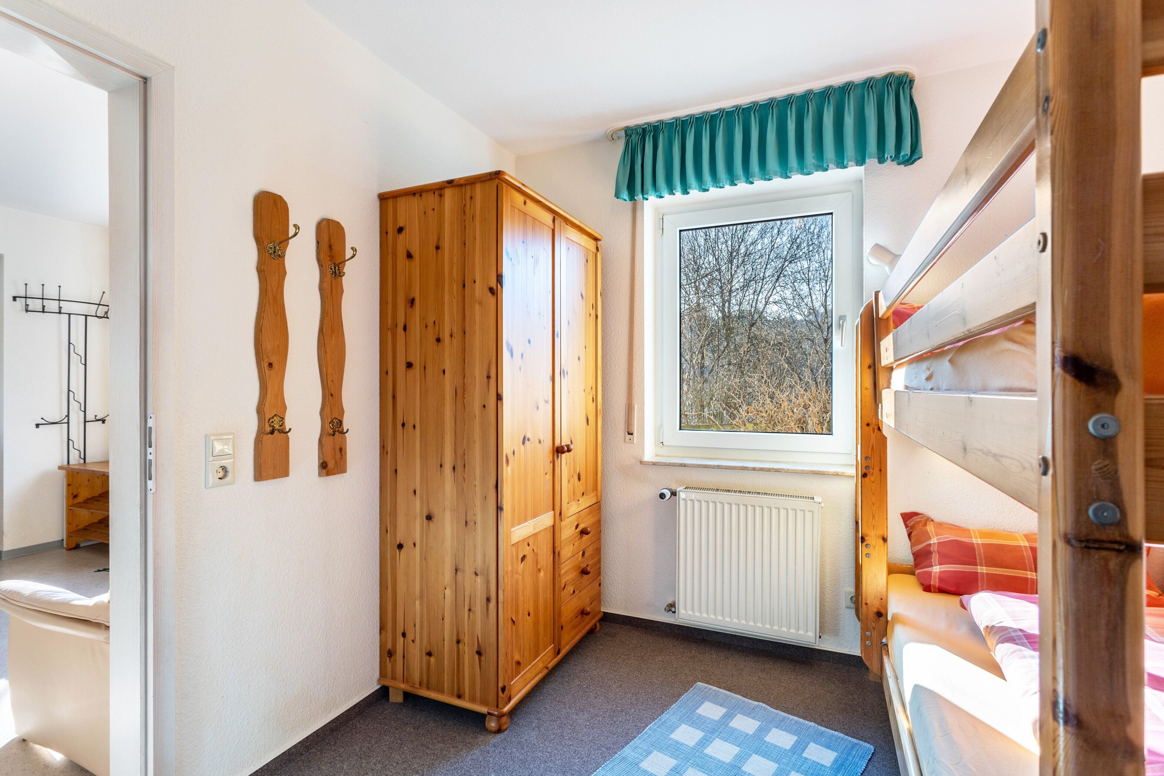 A room with a wooden wardrobe, a window with a view, a radiator, and a bunk bed.