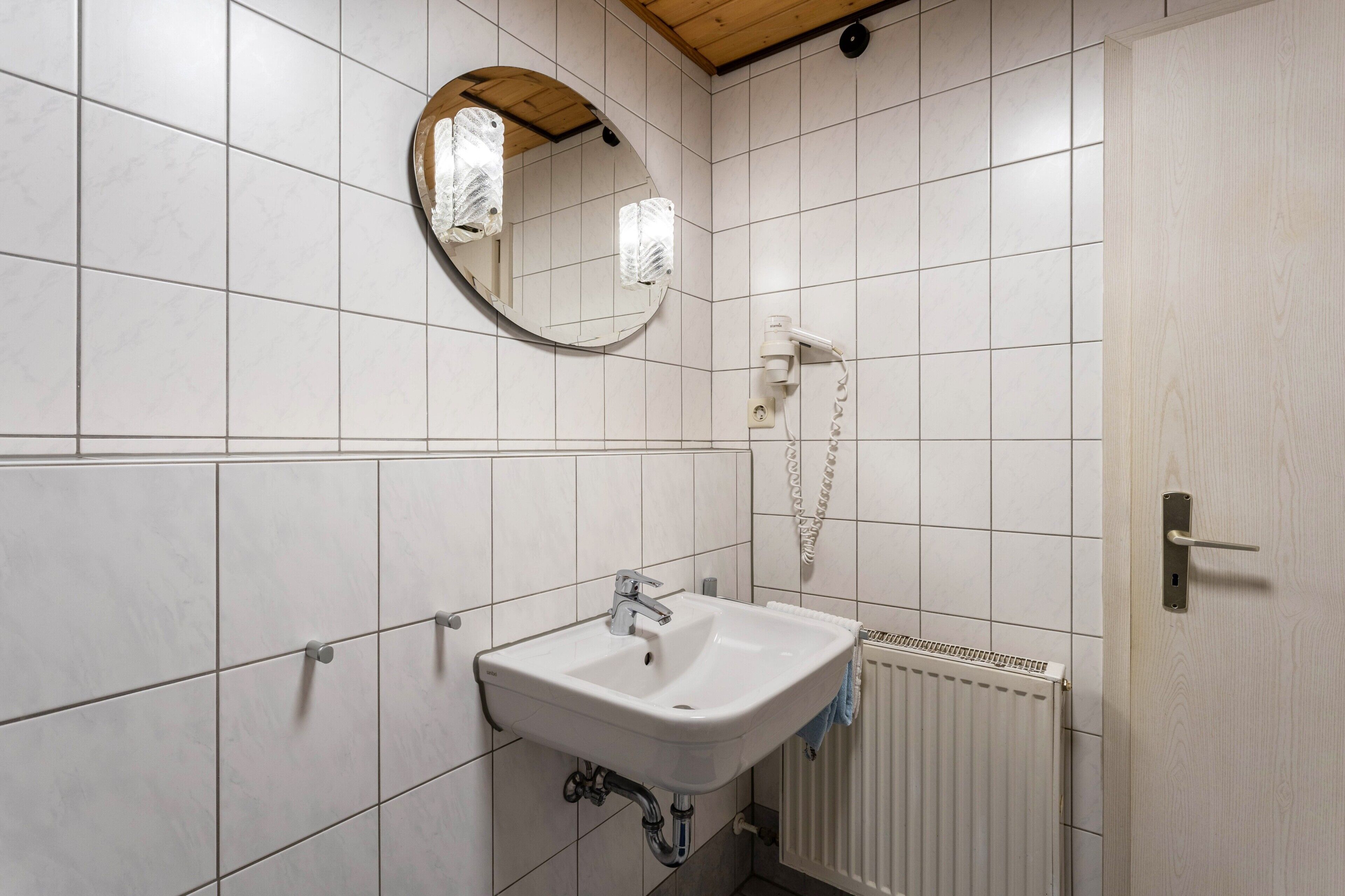 Ein Badezimmer mit weiß gefliesten Wänden, einem runden Spiegel, einem weißen Waschbecken, einem Handtuchhalter und einer Tür mit Holzboden.
