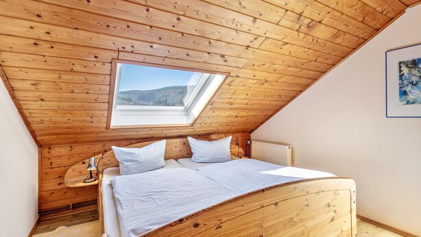 Une chambre avec un plafond en bois incliné, une lucarne, un lit avec une literie blanche et un radiateur.