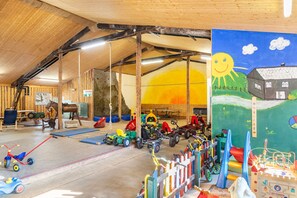 Een ruime binnenspeelruimte met diverse kinderspeelgoed en -apparatuur, een houten plafond en een muurschildering.