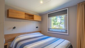 A bedroom with a bed, a window with a view, a wooden shelf, and a wall-mounted light. - Amorosa Mobile Home, Bonne Anse Plage at La Palmyre (La Palmyre)