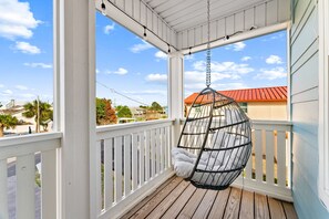 En veranda med en hängstol, vita räcken och utsikt över en byggnad med rött tak.