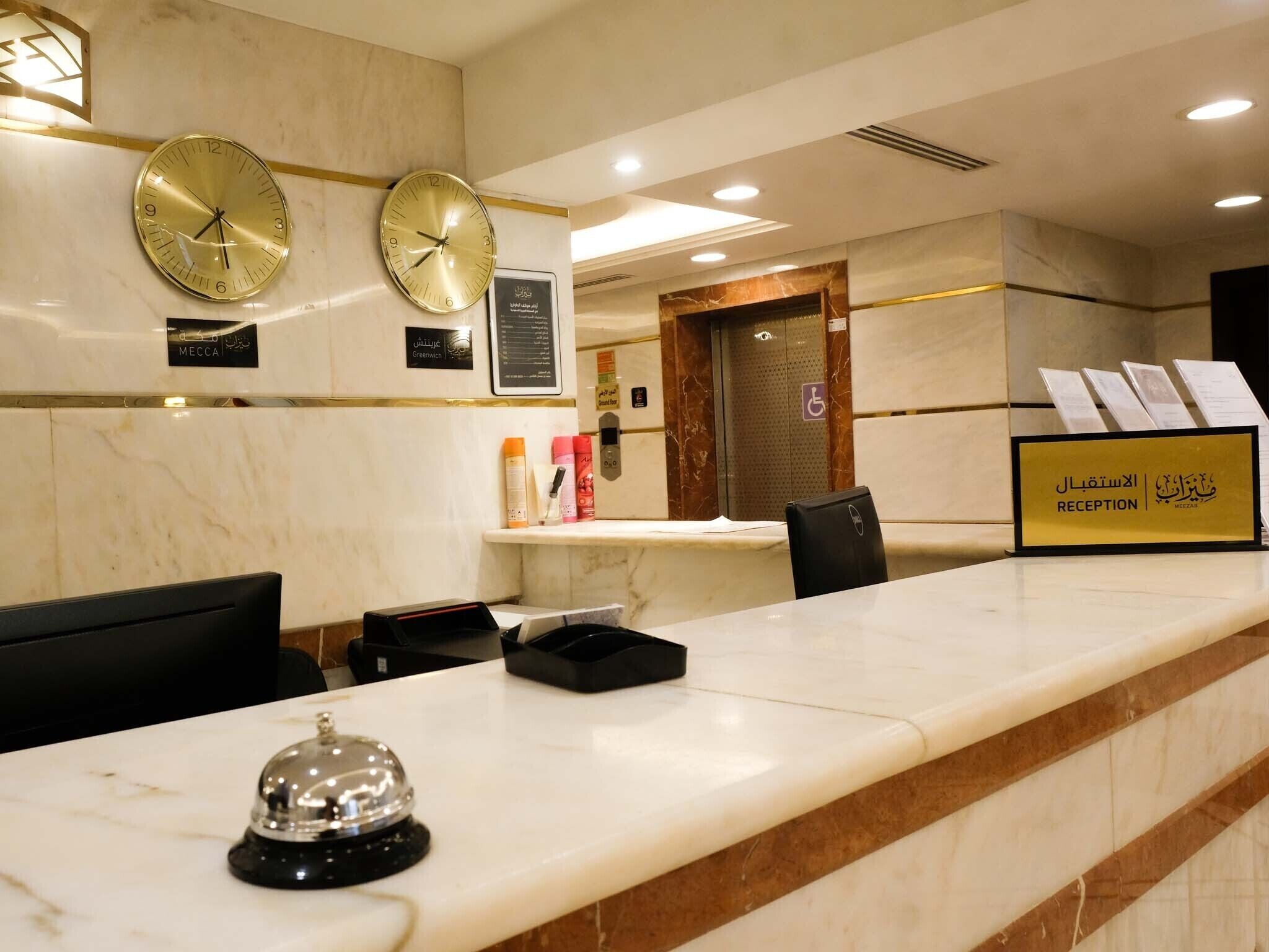 Zona de recepción de un hotel con mostradores de mármol, dos relojes de pared, un mostrador de recepción y un cartel que indica la zona de recepción.
