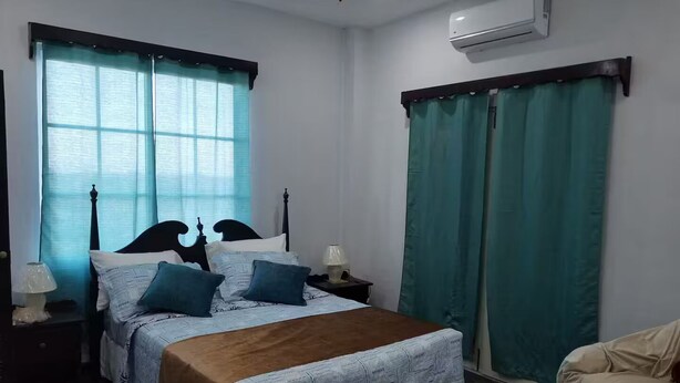 Ein Schlafzimmer mit einem Bett, zwei Nachttischen, einer Lampe, einem Sessel und einem Fenster mit Vorhängen.