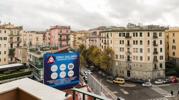A cityscape with multi-story buildings, a prominent billboard advertising DINO GALIANO, and a street with parked cars. - VomeroMetroNest By Dimorra  (Napoli)