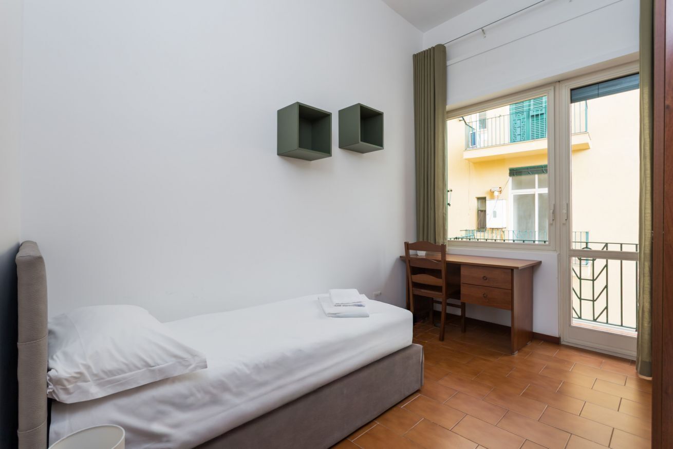 Quarto com cama, escrivaninha e cadeira. Possui varanda com vista para um edifício.