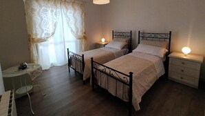 Ein Schlafzimmer mit zwei Betten, einem Nachttisch mit Lampe, einem kleinen Tisch mit Stuhl und einem Fenster mit Vorhängen.