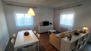 Ein kompakter Wohnraum mit Esstisch, Sofa, Fernseher und einem kleinen Beistelltisch.