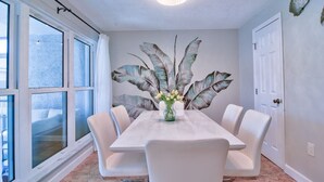 Un comedor con una mesa blanca, seis sillas, un florero con flores, un mural grande con hojas tropicales y una puerta.