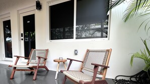 Two wooden chairs with woven seats and backs, a small round table, and a potted plant beside a building with large windows. - Casitas at Coconut Grove (Miami)