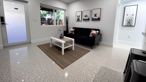 A modern living room with a black sofa, a white coffee table, and a window with a view of greenery. - Casitas at Coconut Grove (Miami)