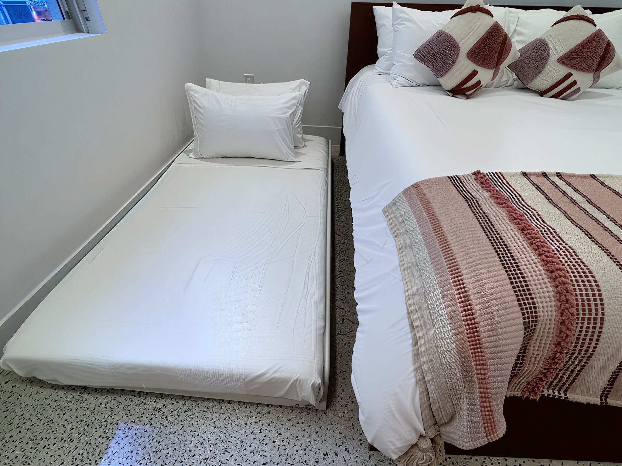 A small, neatly made bed with white bedding and a single pillow, a larger bed with white bedding and multiple pillows, and a patterned blanket.