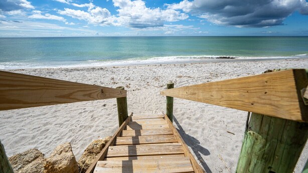 Una escalera de madera que lleva a una playa de arena, con vistas al océano y un cielo parcialmente nublado.