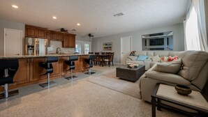 Una cocina moderna con armarios de madera y una isla central, un comedor con sillas, una sala de estar con sofá y televisor, y una isla de cocina con taburetes de bar.
