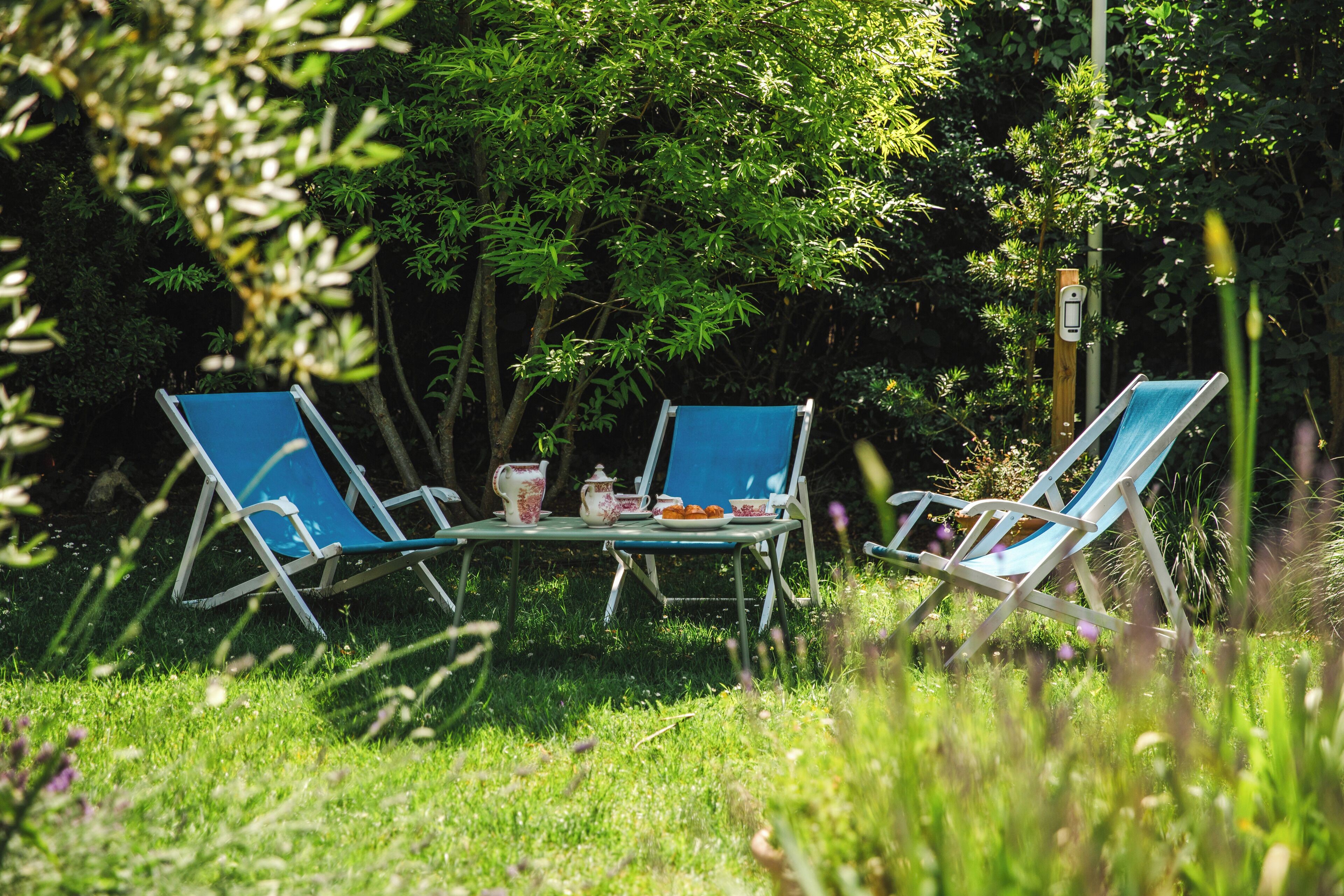 Two blue lawn chairs and a table with a teapot and cups in a garden setting.