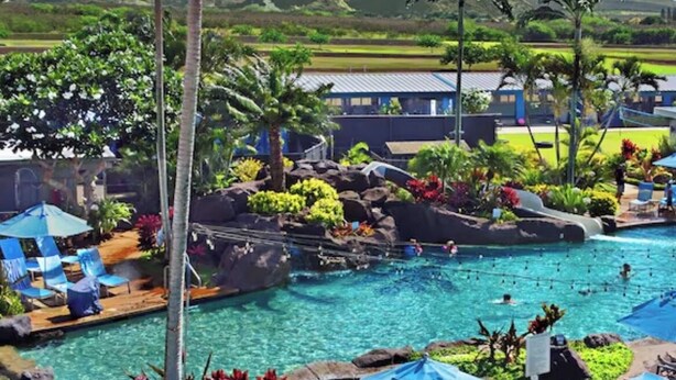 Een zwembadgedeelte van een resort met ligbedden en parasols, omgeven door tropische beplanting en uitzicht op verre heuvels.