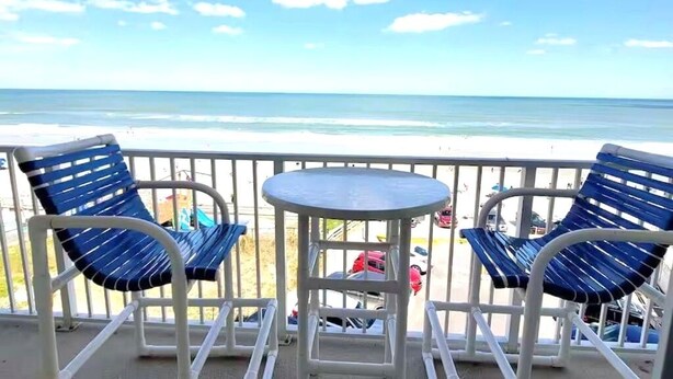 Un balcone con due sedie blu e un tavolino rotondo, con vista su una spiaggia e un cielo sereno.