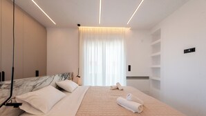 Un dormitorio moderno con cabecera de mármol, una cama grande con colcha color crema, dos toallas enrolladas y un ventanal con cortinas translúcidas.