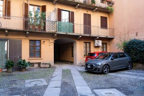 Un bâtiment à deux étages avec une cour intérieure. On y trouve des plantes en pot et une voiture rouge garée devant.