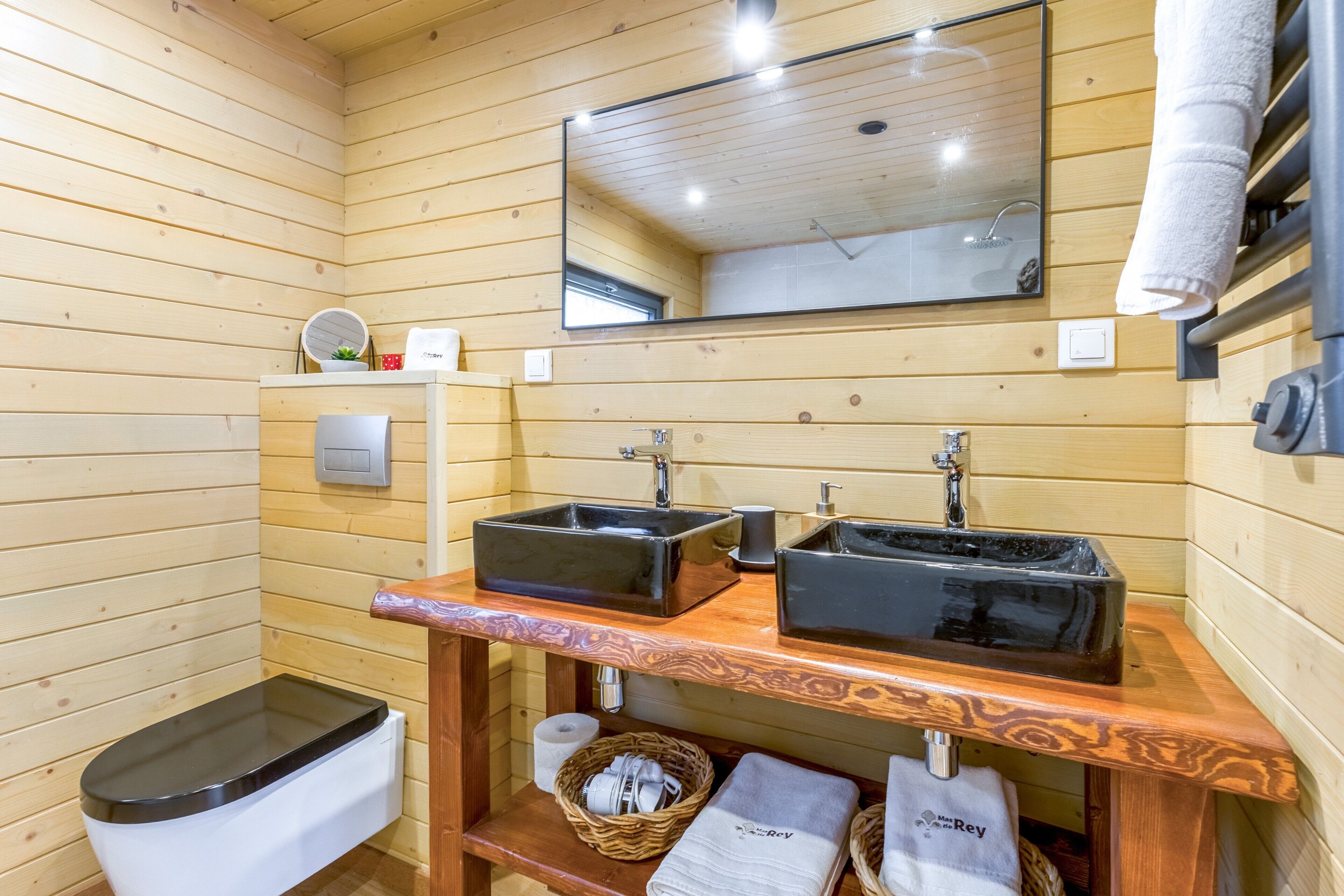 Une salle de bain aux murs lambrissés, avec deux lavabos noirs, un miroir, des toilettes et une étagère où sont posées des serviettes.