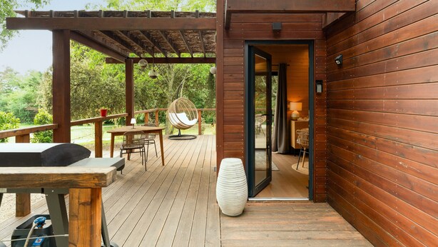 Una terraza de madera con pérgola, una mesa, sillas y un sillón colgante.