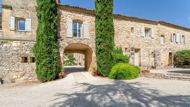 Un bâtiment en pierre doté d’une arche centrale, de grands pots de plantes et d’une allée gravillonnée.