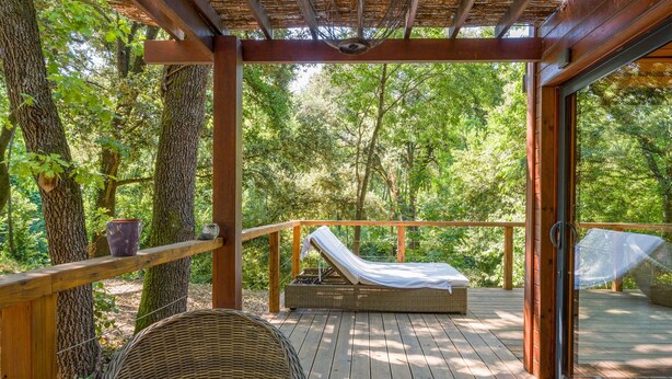 Un pontile in legno con una poltrona da relax e un tavolo, immerso nel verde degli alberi.