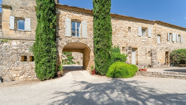 Un bâtiment en pierre doté d’une arche centrale, de grands pots de plantes et d’une allée gravillonnée.