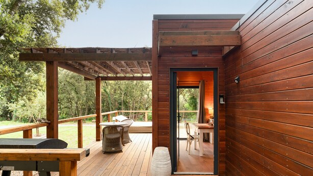 Una terraza de madera con mobiliario exterior, una pérgola y una puerta que da a un espacio interior.