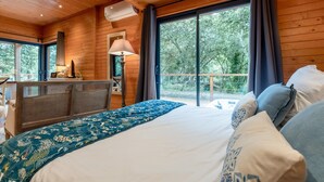 Un dormitorio de cabaña de madera con cama, lámpara de noche y vistas a árboles a través de amplios ventanales.