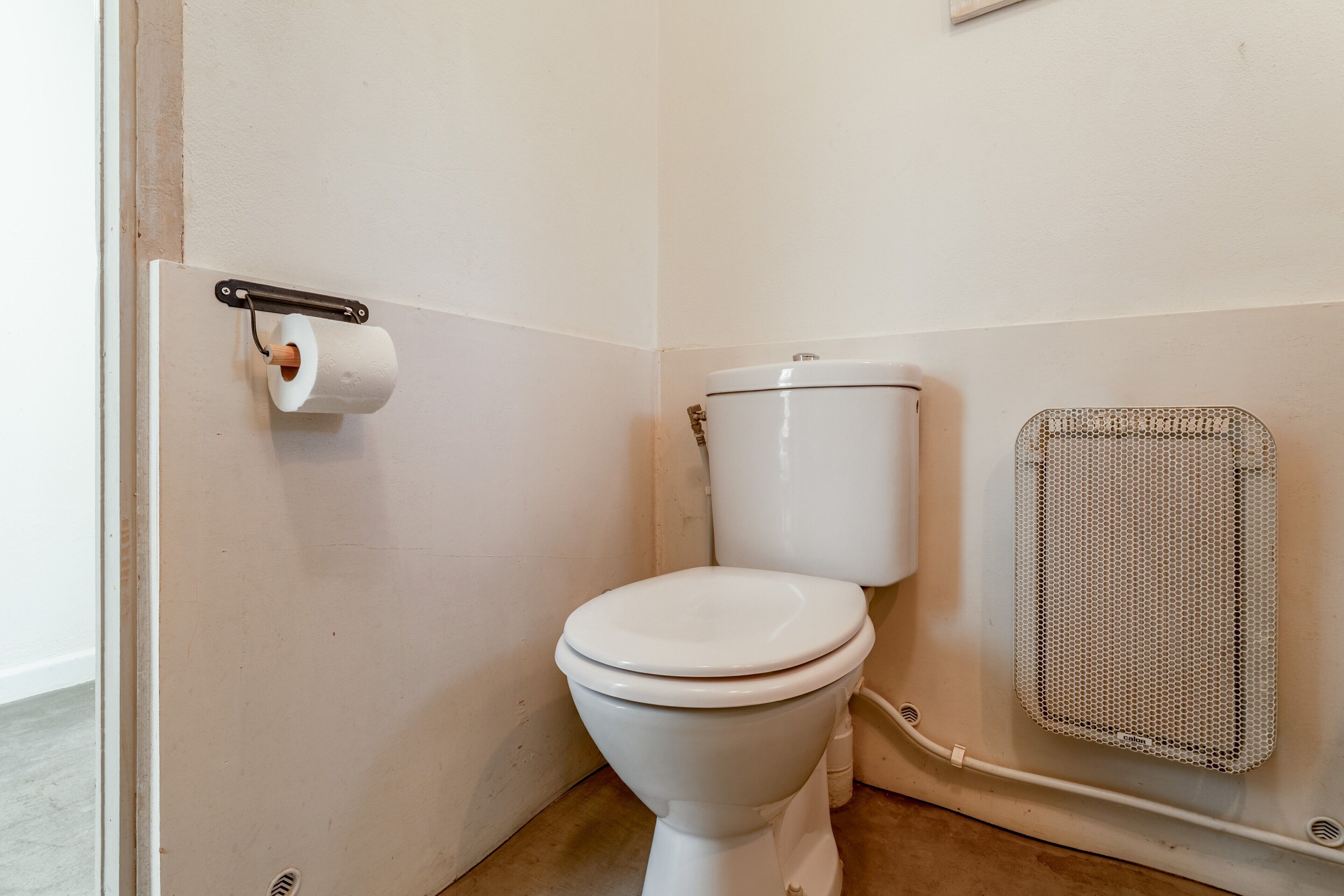 A white toilet with a paper towel holder, a metal vent, and a white wall.