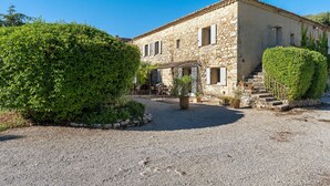 Un bâtiment en pierre avec une allée gravillonnée, un grand buisson et un escalier menant à l’entrée.
