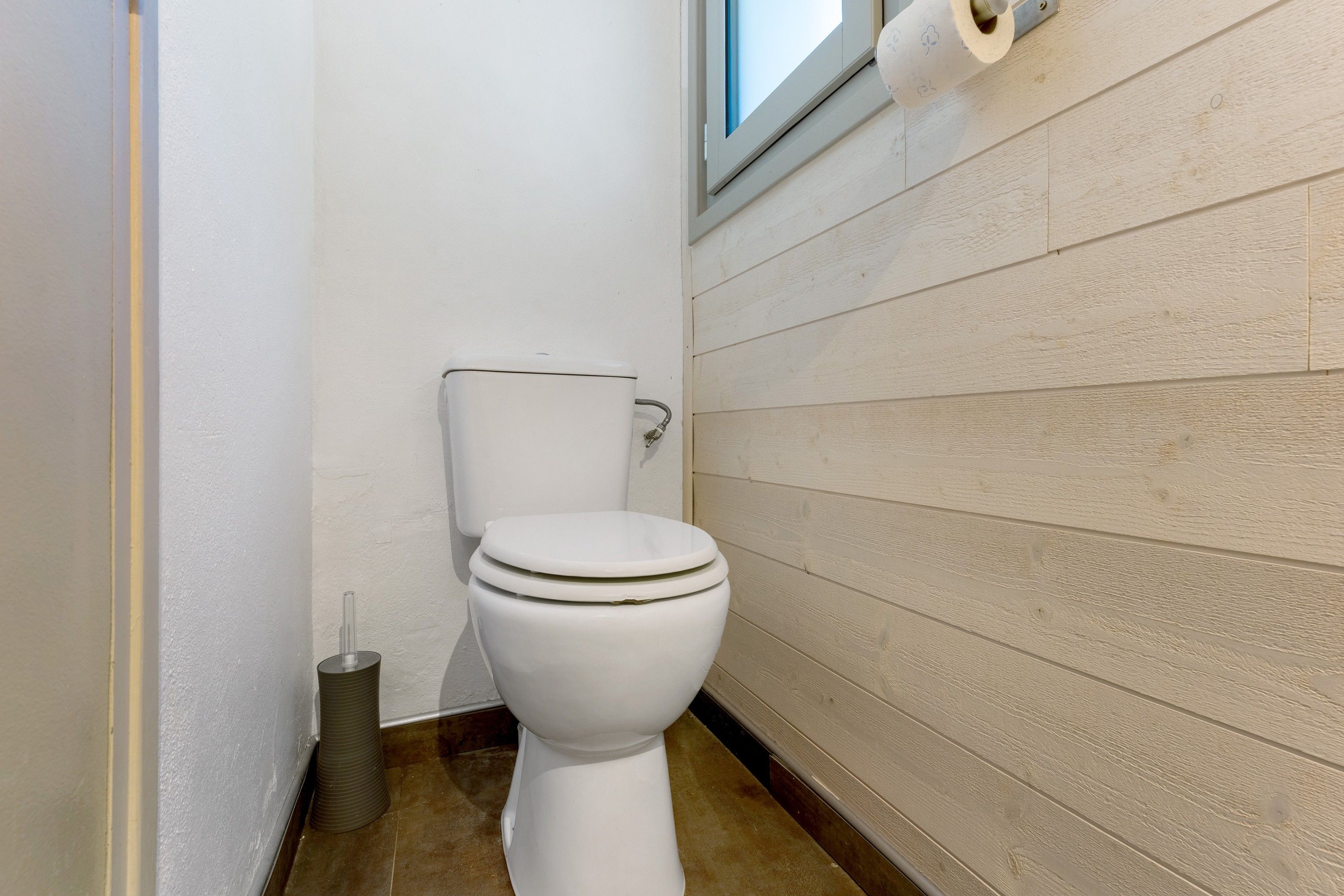 A small, white toilet with a tank and lid, positioned against a wall with horizontal wooden panels.