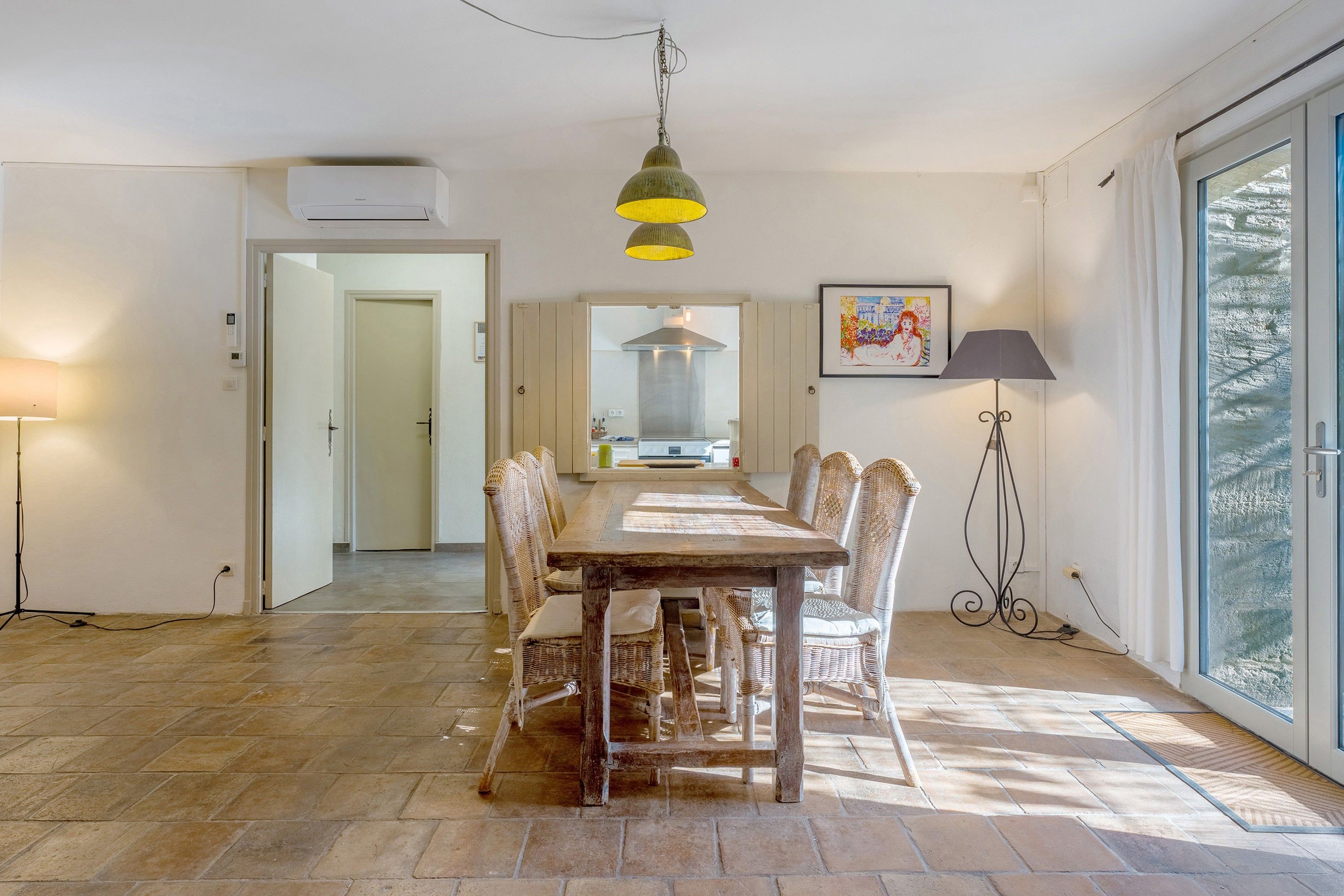 Een eetgedeelte met een houten tafel en rieten stoelen, een hanglamp en uitzicht door een glazen deur.
