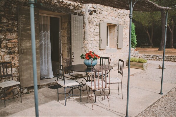 Un espace extérieur aménagé pour les repas, avec un bâtiment en pierre, des chaises en métal, une table ronde et un vase rempli de fleurs.