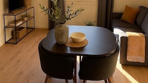 Una sala de estar moderna con una mesa redonda, dos sillas, un sofá, un televisor sobre un mueble bajo y una ventana con cortinas.
