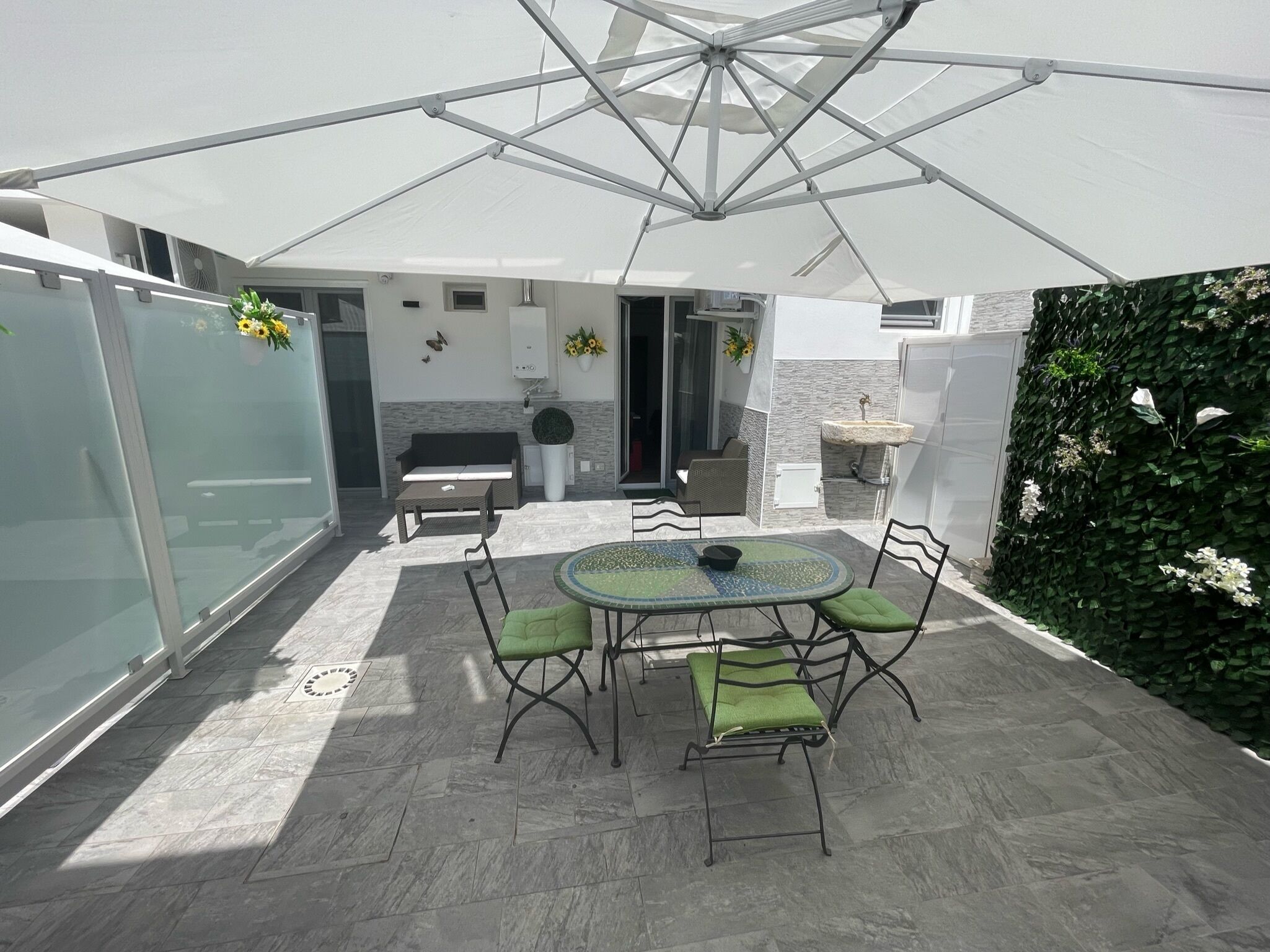 Eine Terrasse mit rundem Tisch und Stühlen, einem weißen Sonnenschirm und einer grünen Wand mit Kletterpflanzen.