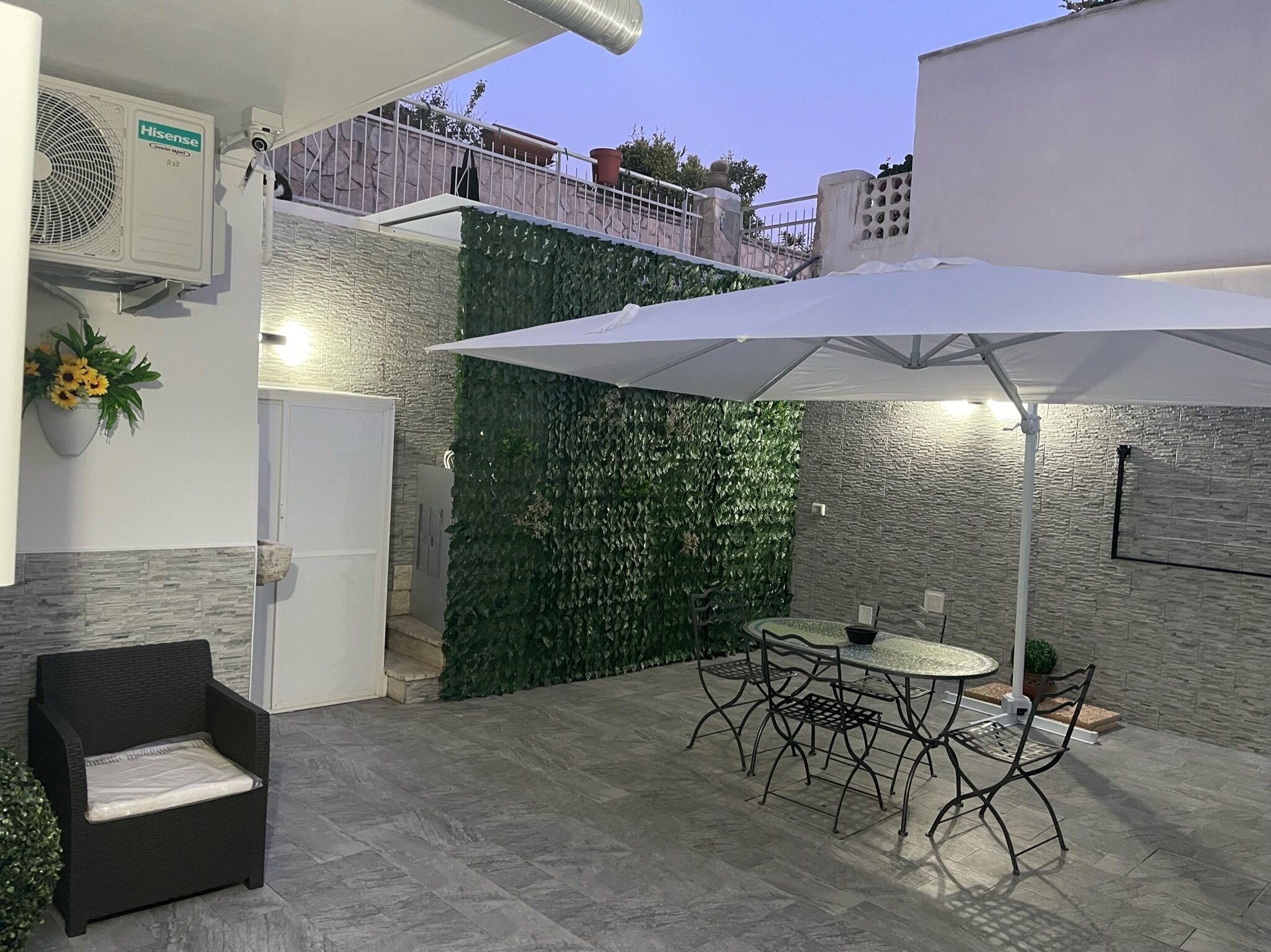 An outdoor patio with a table and chairs, a white umbrella, and a wall with green foliage.