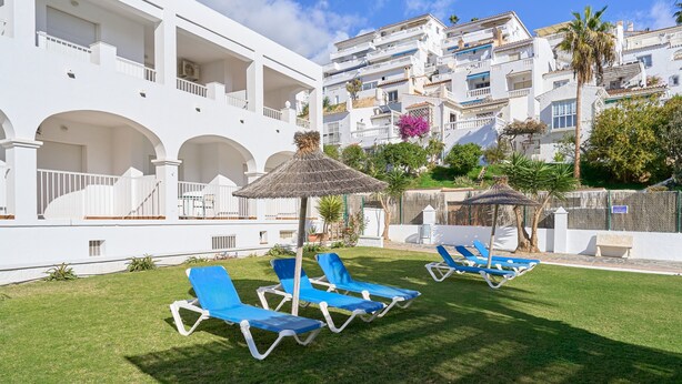 En vit byggnad med flera balkonger, en gräsplätt med blå solstolar och ett palmträd.