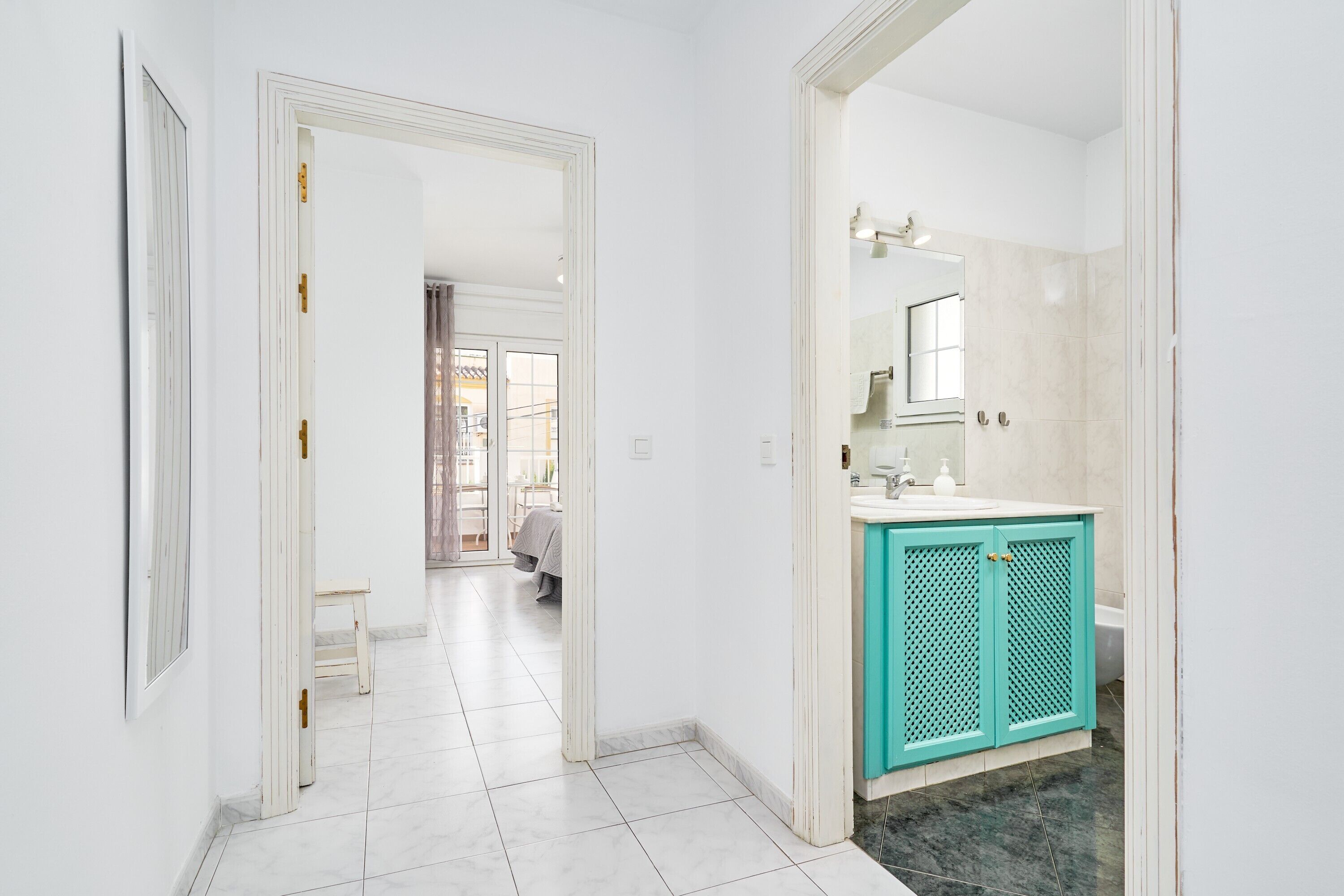 Een gang die leidt naar een badkamer met een witte deur, een bankje en een blauwgroene kast.