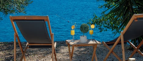 Two wooden lounge chairs facing a body of water, with a small table between them holding a potted plant and two glasses of orange juice.