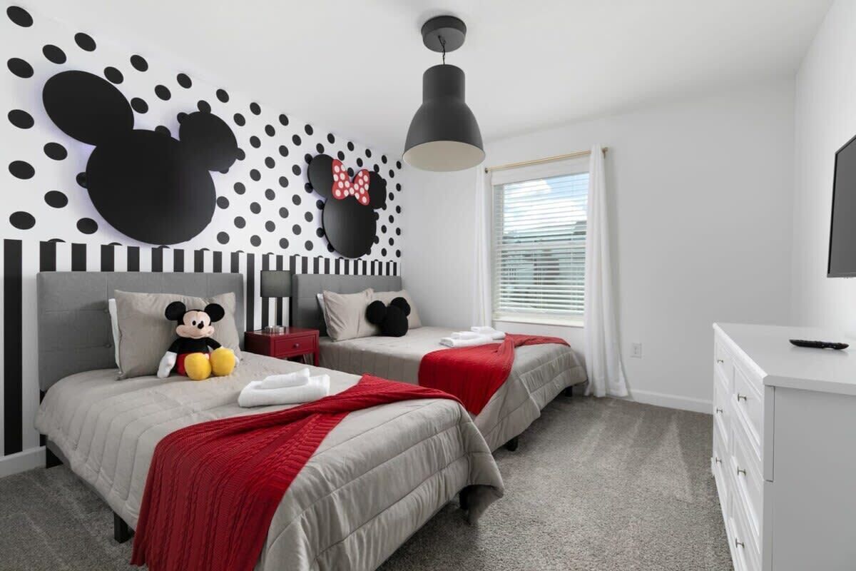 A bedroom with two beds, a Mickey Mouse wall decoration, a red bow, and a ceiling light.