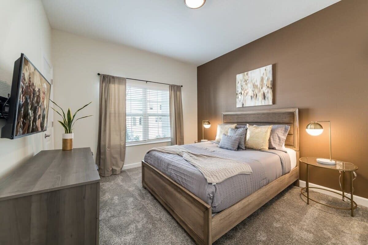 A bedroom with a bed, bedside tables, a desk, a window with curtains, and wall art.