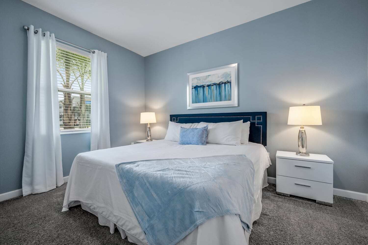 A bedroom with a bed, two bedside tables, a window with curtains, and a painting on the wall.