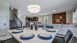 Uma sala de jantar moderna com uma mesa branca, cadeiras escuras e uma luminária central.