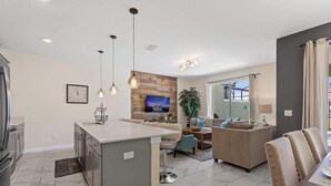 A modern kitchen with a central island, a dining area with a table and chairs, and a living room with a sofa and TV. - Amazing Resort Near Disney 4BR Townhome (Kissimmee)