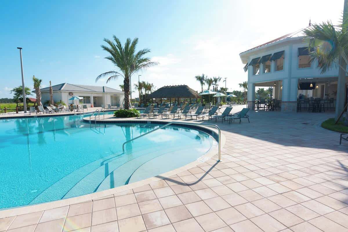 Une piscine entourée de palmiers et d’un bâtiment doté d’une zone couverte, avec des chaises longues et des parasols à proximité.