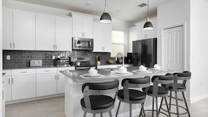 A modern kitchen with white cabinets, a central island with bar stools, a microwave, and a refrigerator. - Chic & Spacious Best Vacation Home (Kissimmee)