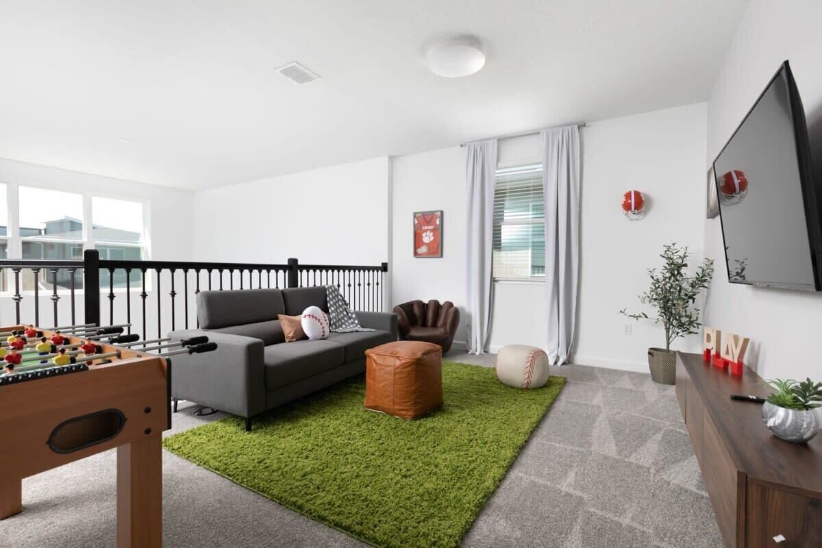 A modern living room with a foosball table, a grey sofa, a brown leather armchair, a green rug, a wooden cabinet, and a TV on the wall.